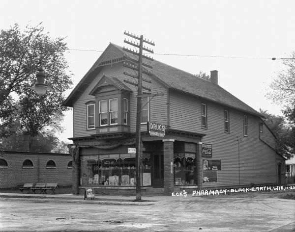 Front and side of the two-story drug store on the corner of the street. The store advertises Kodaks, ice cream, soda, cigars, Coca-Cola, Victor records, and cigarettes. There are three benches on the left of the store and a large utility pole is in front.