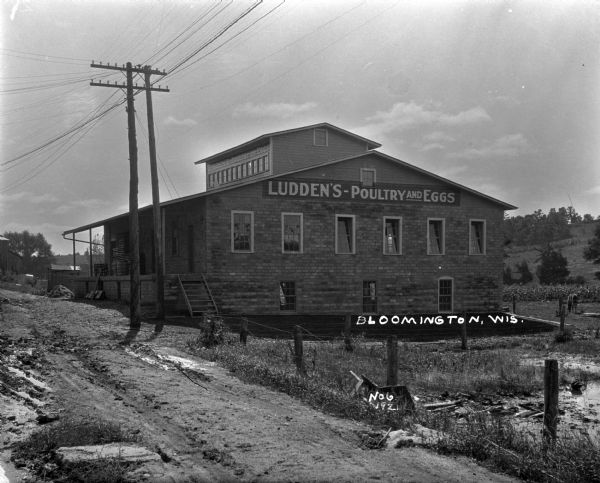 Exterior view of Ludden's Poultry and Eggs from the dirt road. The shop features a porch, gable roof, and fenced yard. There are wooden crates and barrels on the porch.