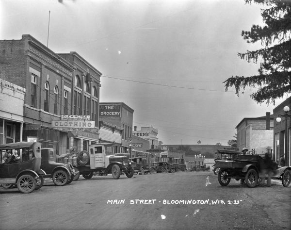 Rows of cars are parked at shops on Main Street. The shop signs (from left to right) are: Doctor R.H. Merrifield Dentist, Clem Meyer — Dry Goods and Groceries, Clothing — Shoes for the Whole Family, Woodhouse and Bartley, Groceries and Meat, Ludden's Store for Men — Clothing — Furnishings — Hats — Shoes, Oates and Ellis Hatchery, and Sprague's Recreation Parlor.