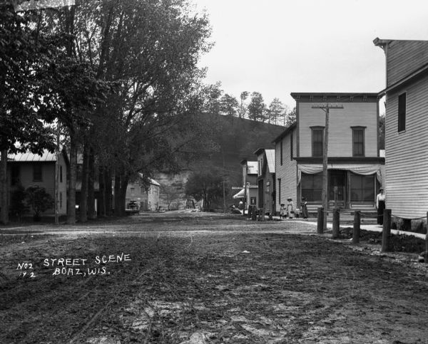 View down dirt street. Two women and three children pose at the intersection. Farther down the street is a post office. There is a bridge at the end of the road and a steep hill in the far background.