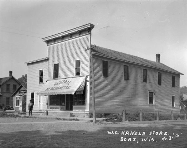 Exterior of the "General Merchandise - A.C. Hanold groceries" and the "garage and auto livery." A man stands outside the store.
