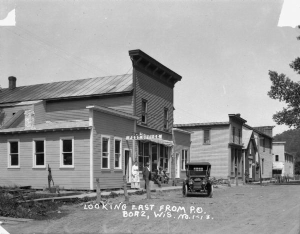 Exterior of the post office. On the sidewalk, a man and woman stand and three men sit on the steps. A black car is parked at the curb. A hand-powered pump is on the side of the building.