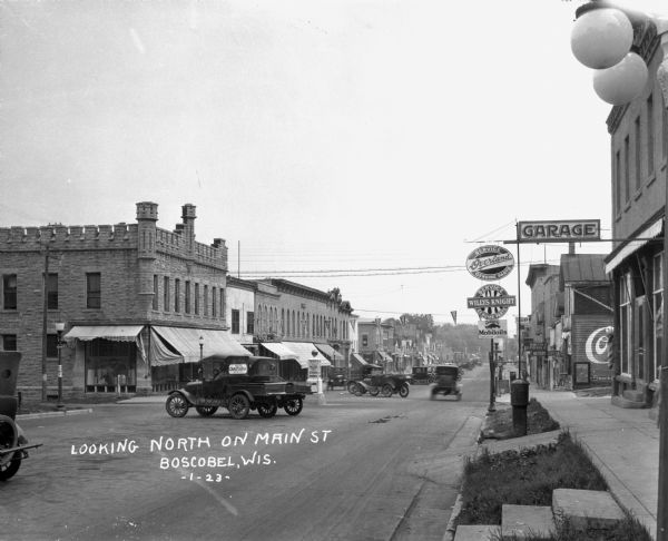 A busy street scene on Main Street and Wisconsin Avenue. Cars are parked in the middle of the street, and triangle flag banners hang  above. The shops have signs that say: Ice Cream - Candy - Cigars, Hotel, Smith Auto Sales, and Garage. There are advertisements for: Miller Hardware Company, Chautauqua, Mobiloils, Willys-Knight, Overland, and Coca-Cola.