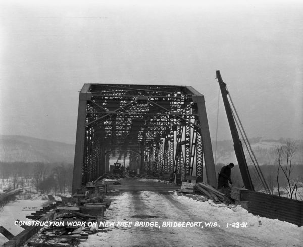 The entrance to the bridge during construction. A worker works on the side of the bridge on the right. Other workers in the middle of the bridge lay boards. There is a sign that reads, "loitering prohibited."
