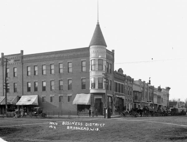A main street intersection with many stores and shops. The stores include the Bank of Brodhead and A.J. Wagner's drugstore. Three boys stand at the corner. Cars and horses are parked along the curbs.