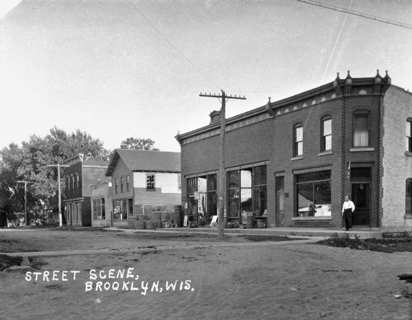 View across an intersection of a man standing at the corner entrance of an untitled shop. Down the sidewalk on the left a young boy leans over to talk with another boy sitting on the steps of a store with display windows. There are rolls of fencing mesh stacked in an alley along the side of the store.