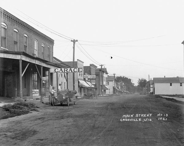 View down middle of commercial street. On the left, two men pose by a car at an automobile repair shop. The shop advertises Polarine motor oil and Willard Storage Batteries. Next door is George Foehringer's shoe store.