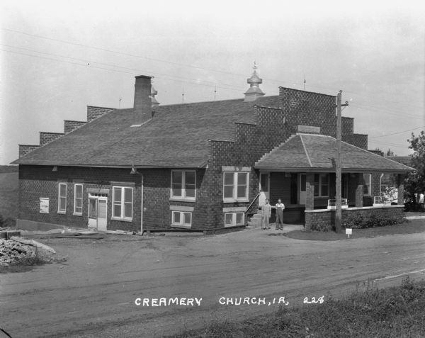 View across road of two men posing outside a creamery. The creamery has a roofed loading dock on the side.