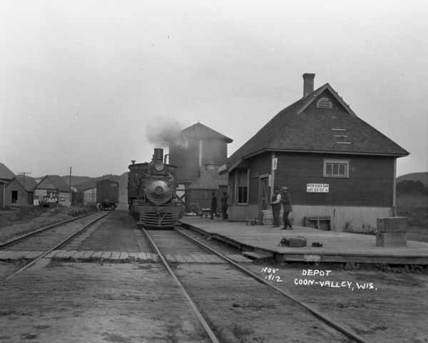 Train at Coon Valley depot. Several men stand on the platform. The depot has a water tank. There are advertisements for Pearl White Soap, the inter-state fair, and 5 cent cigars.