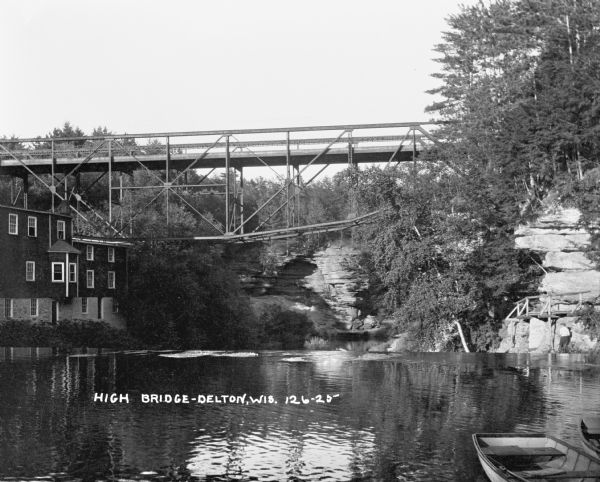 View across water of a deck truss bridge over the dam at Mirror Lake. Timme Mill is on the left side of the lake. There are boats in the foreground, and the shorelines are steep, rocky cliffs covered with trees and plants. A man or young boy is standing on the right just under a wooden footbridge.