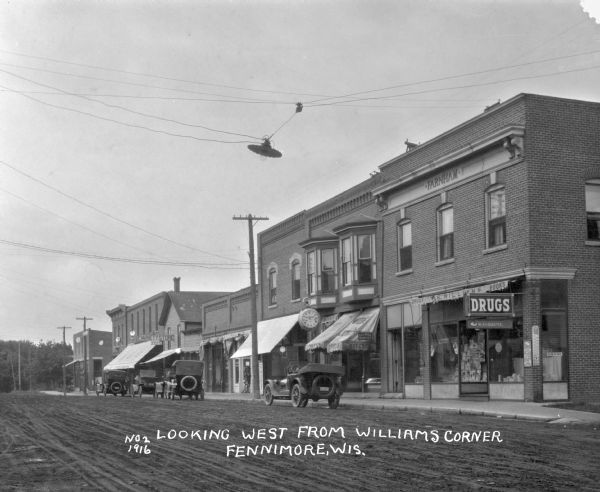 View across street of a row of stores, including R.S. Williams drugstore, C.C. Lewis's dental office, Weber's watch and jewelry store, and a variety store. Automobile are parked along the curb.