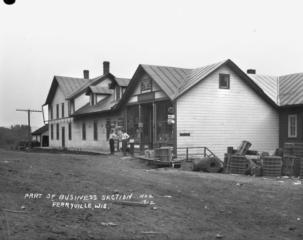 View across road of two men, a dog and a boy standing in front of a variety store.