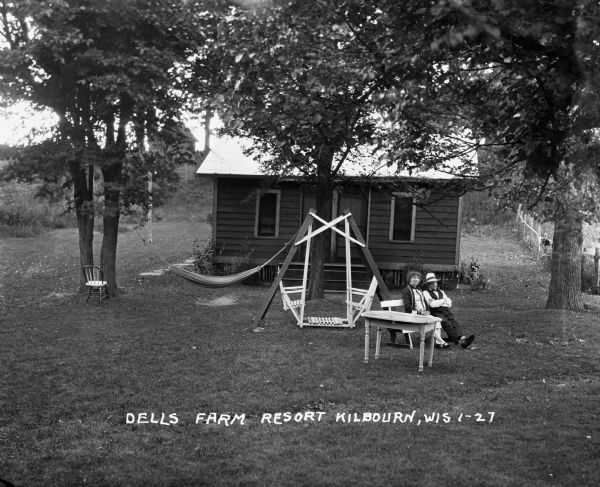 A man and woman sit on a bench outside a cabin at the Dells Farm Resort. A chair, hammock, lawn swing and table are on the lawn.