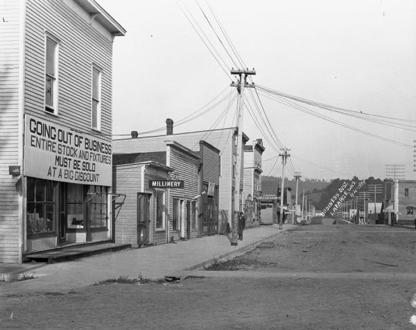 View down a street in La Farge. A store's sign reads, "Going out of Business - Entire stock and fixtures must be sold at a big discount." Other shops include a millinery, restaurant, grocery store and shoe store.
