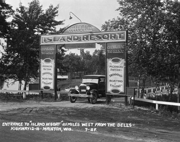 Car parked underneath the entrance gate to the Island Resort at the Wisconsin Dells, 25 miles from the Dells on Highway 12-16 near Mauston. Entrance gate advertises swimming, boating, fishing, picnics, and parties.