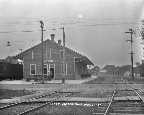 Depot View down railroad tracks of the exterior of the Mazomanie train depot. Two sets of railroad tracks are crossing a dirt road in the foreground, and a railroad car is sitting on the tracks on the other side of the depot.
