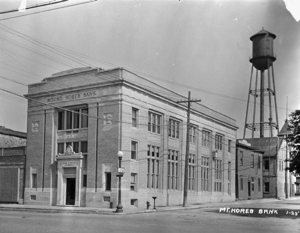 View from intersection of the exterior of the three-story Mount Horeb Bank, next door to the printing office. There is a water tower in the background.