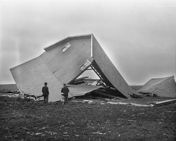 Two men and a child look at a barn that has collapsed, possibily due to a windstorm or tornado.