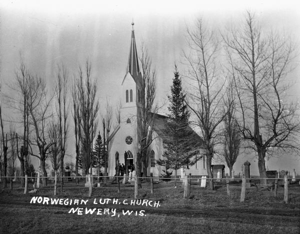 View across lawn towards Norwegian Lutheran Church and cemetery. Well-dressed men and women stand at the entrance to the building. There are graves behind a fence in the foreground.