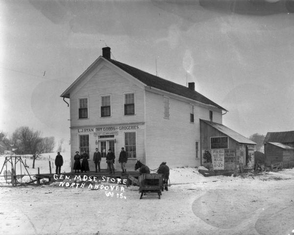 Six people standing on the porch of L.J. Ryan Dry Goods and Groceries. A sign above the door says: Post Office North Andover." A team of two horses are pulling a sled. There is a lawn swing to the left of the porch of the store. On the right are posters and advertisements on a lean-to addition on the side of the building.