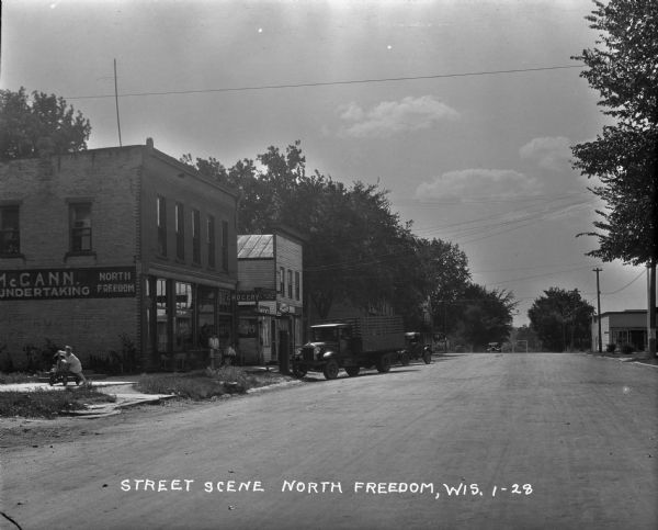 View down street. On the left is the McCann. Undertaking building and grocery store. Two children play on a wagon on the sidewalk. Five people are gathered in front of the storefront, and a man sits in one of two trucks near a Pennco Gasoline pump.