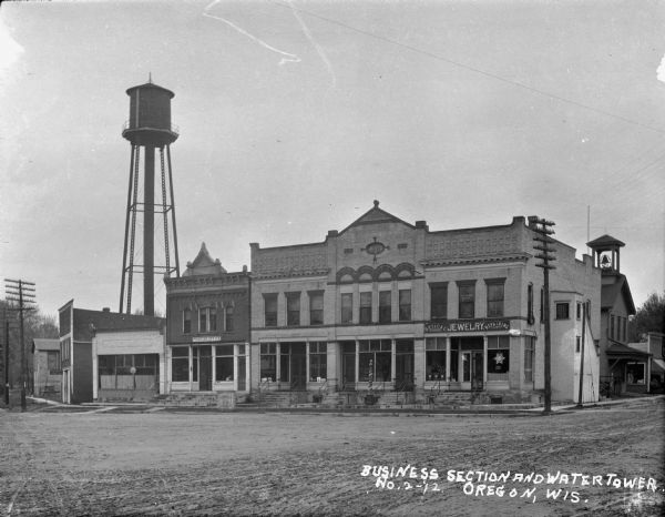 View across road of the central business section. The commerical buildings along the sidewalk include a printer, butcher, barber and jeweler. Above the shops are offices. The year "1898" is marked on the upper center of the facade above the office windows. On the right just behind the shops is a building with a bell tower, and rising high above and behind the storefronts is a water tower on the left. In front of the meat market on the sidewalk is a scale with the words "Howe U.S. Standard."