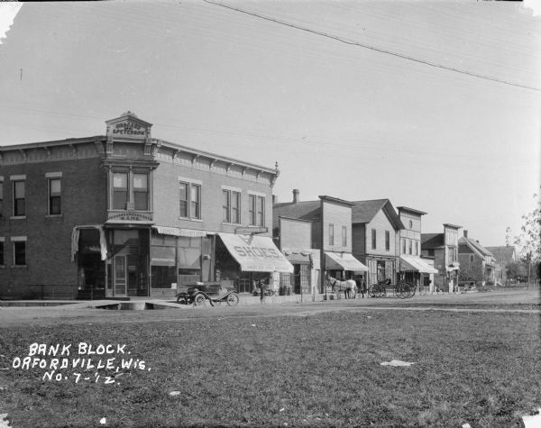 View across lawn of a commercial block, including the Farmers Merchants Bank, general store, and meat market. Horses and wagons, and an automobile are parked along the curb.