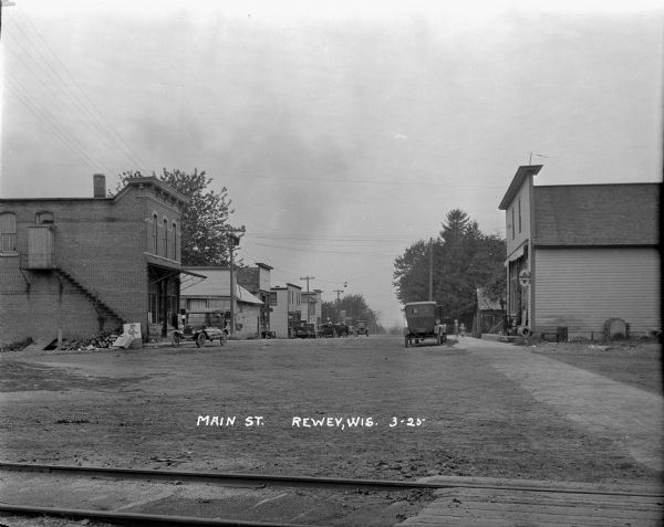 View across railroad tracks of Main Street. Commercial buildings are on both sides of the street, and cars are parked along the curb. A service garage in the far background. Two children are walking along the sidewalk on the right.