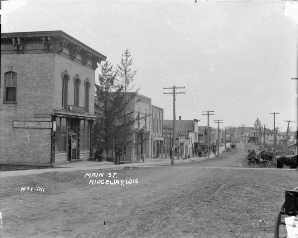 A downhill view of Main Street with a shoe store and other buildings on the left. Some homes and a church in the distance. Horses and wagons in the street and a few pedestrians.