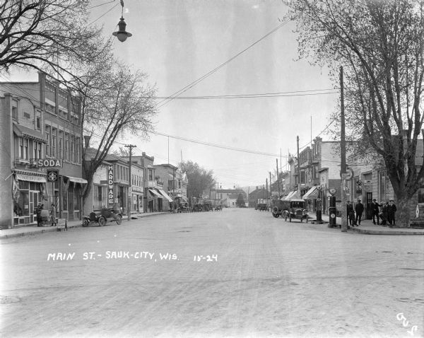 View down center of Main Street. On the left is a soda shop/confectioner, with two women and a baby carriage on the sidewalk in front. Further down is a chiropractor's office, hardware store, bakery and two restaurants. On the right is a hotel, drug store, and a gas station at the corner in the foreground, with a group of men and boys standing in front. Cars are parked along both sides of the street.
