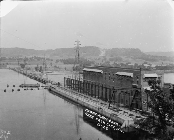 Elevated view of the power plant and dam on the Wisconsin River. Two workers are walking near the center. On the opposite shoreline are fields and tree-covered hills.