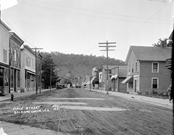 View down sidewalk of Main Street with commercial buildings on both sides. A steep hill with trees is in the far background. On the left are doctor's offices, and further down parked cars along the curb, with pedestrians. On the right are a billiard hall, shoe repair shop and the Kickapoo Scout building. On the hill in the background are houses and barns.
