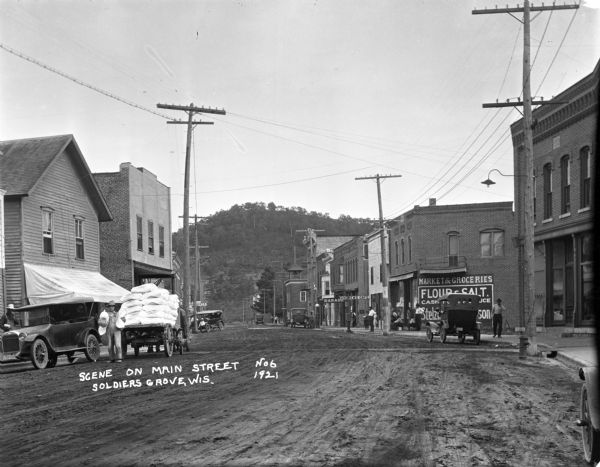 View down Main Street with steep, tree-covered hill in the far background. On the left a man is holding flour sacks and standing near a horse-drawn wagon loaded with more sacks of flour. Next to him is an automobile parked at the curb with another man standing on the sidewalk. On the right side of the street is a grocery store, cafe, and service garage, and further down is a brick building with a bell tower. There are cars parked along the curb and a number of pedestrians.