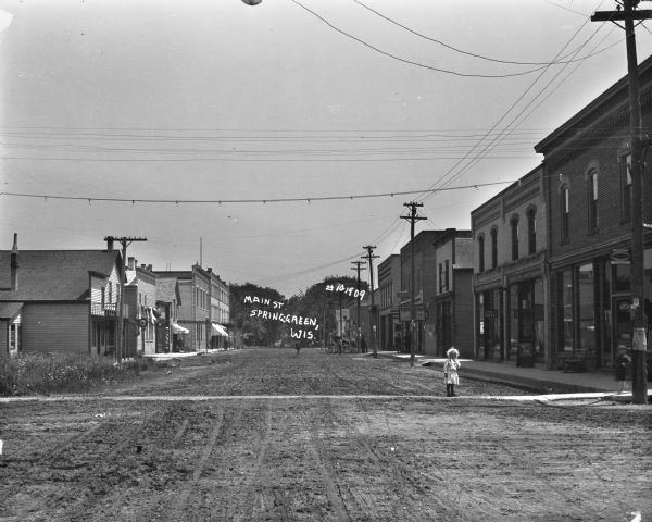 View down center of Main Street which is unpaved. A young girl wearing a bonnet is standing in the road on the right, near another child standing at the corner. A group of men stand near a ten cent store in the background on the right. On the left side of the street is a barbershop, restaurant and livery. Horses and wagons are parked along the curbs.