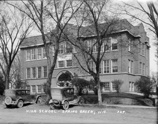 Spring Green High School Exterior view from road of the high school, a three-story brick building with an arched doorway and trees and bushes in the lawn in front. Two cars are parked in front.