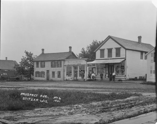 View of Prospect Avenue. A group of men and a woman standing in front of the store, which is a two-story wooden building with shuttered windows, next to a restaurant/soda fountain and general store. Another building with shuttered windows is on the right.