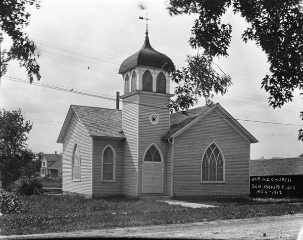 View from road of the German Methodist Episcopal Church. The double doorway has an arched window above it, and another circular window with a decorative frame. The bell tower has shutters in a dome with a weather vane. There is a stables on the right.