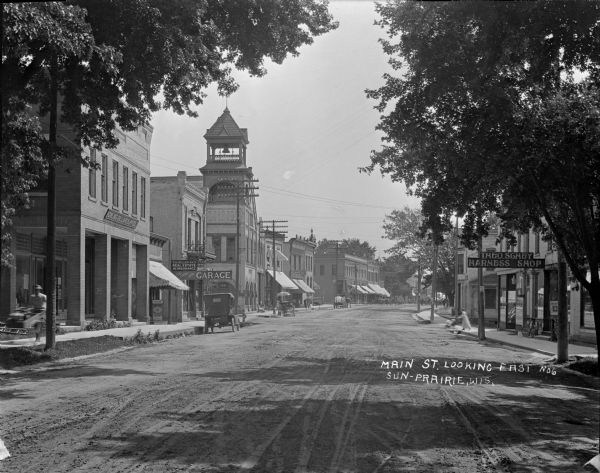 View down Main Street. On the left is a bowling alley/billiard parlor, a real estate/insurance business, and a livery/garage. On the next block is City Hall which has a bell tower. There is a harness shop on the right with a bicycle leaning up against the window. A man is pushing a cart along the sidewalk on the left, and a woman is pushing a stroller on the right. There is an automobile and horses and delivery wagons along the street.