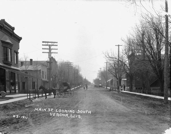 View down center of Main Street. On the left is a storefront with large display windows and a barrel near the entrance. In front is a horse and buggy hitched up to a post. A man with a newspaper is standing on the sidewalk, and another man is leading his horse down the street. On the right is a couple walking with their infant.