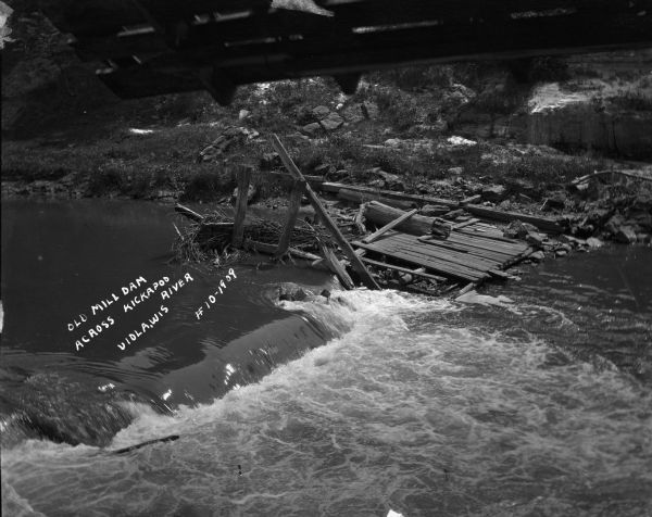 View looking down at the remains of an old dam. Driftwood has been caught up at the broken wood pilings on the far shoreline. The river is flowing fast, and in the foreground at the top is what may be part of a bridge.