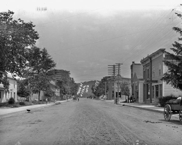View down center of Main Street, with a feed store and other businesses on the left. A grocery store, a barbershop and other businesses are on the right. A group of men are gathered around the stoop of the barbershop which is on the corner. Further down the street are men with horses. In the far background is a tree-lined hill.