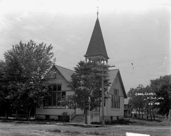 View across unpaved road of the Congregational Church. The wooden, one-story church has a weather vane above the belfry, which has a tall, roofed peak. There is a lamppost at the foot of the stairs to the entrance. Houses are in the background.