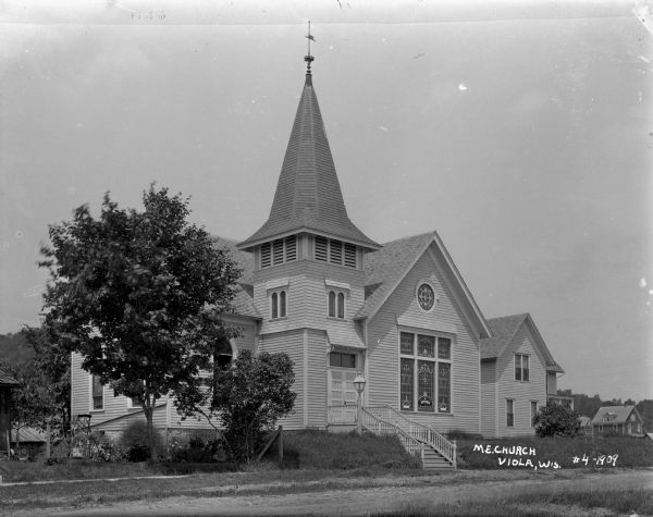 View across unpaved street of front and left side of the M.E. Church. Stained glass windows are in the front of the church near the entrance, which has a flight of stairs and a lamppost. There is a weather vane on the pointed roof above the belfry. Houses are in the background.