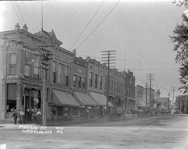 View from intersection of businesses along the left side of Monroe Street. A group of men are standing at the door of the drugstore on the corner, which has an awning that reads: "E.D. Whipple, Druggist." A woman is standing in the doorway of Wendts Restaurant, and a young boy or man stands on the sidewalk. Horses and buggies and wagons are parked along the curb. Other businesses include an attorney, a notary public and a shoe store. Light bulbs are strung up and across the power lines on the left to form what appears to be an arch.