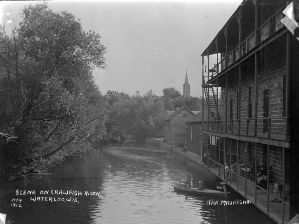 Elevated view of the river with trees on the left. On the right, two young boys are fishing from a rowboat in the river near a landing at the back of a three-story building. The building has a porch at each level, with a roof over the third story porch. Two children are standing near the steps that lead down to the boat. A woman sits on a porch swing nearby. Also on the porch are two taxidermy specimens. In the background above the trees is the steeple of a church.
