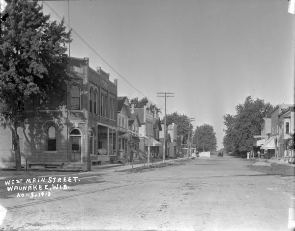 View from intersection down center of West Main Street. On the corner on the left is a brick bank building. A paint store, a shoe store and other businesses are further down along the block. There is some construction going on at a building just before the meat market. A barber, furniture store and a harness shop are on the right. Caption reads: "West Main Street, Waunakee, Wis."