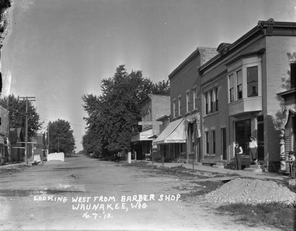 View down Main Street of businesses along the right side. A young boy stands with a shovel near a pile of gravel near the barbershop, which has a barber's pole in front. Two men and a dog stand in front of the brick building next door. Further down is the Farmers State Bank, a furniture store, harness shop, and the "J. Buhlman The Clothier" store. On the left side of the street, a man on a ladder is working on a building. Piles of building materials are in the street.