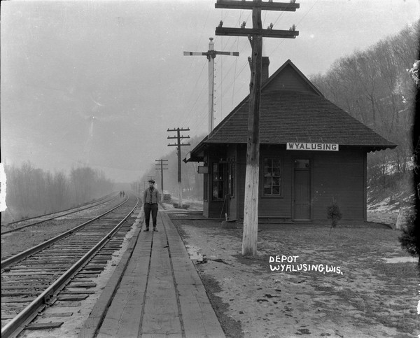 Depot View down platform and two sets of railroad tracks towards depot. One man stands on the platform in the foreground. Two people are further down the tracks walking towards the depot. There is a tall train order signal behind the depot. Snow on the hillside on the right.