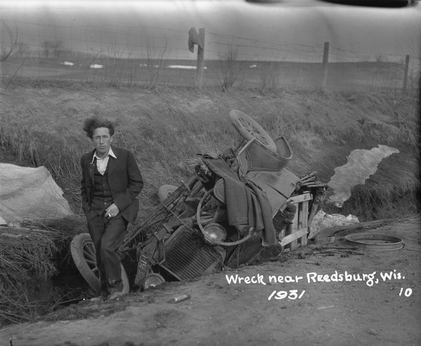 Wreck Near Reedsburg View from road of a man standing near his overturned car smoking a cigarette. His coat is lying across the side of the car. In the background is a barbed wire fence and field.