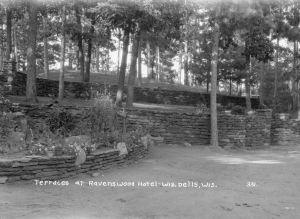 Stone terraces at the Ravenswood Hotel. In the foreground are flowers, some potted, in a raised bed. Two more stone walls are on the hill in the background, with trees on the lawn.